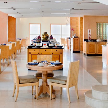 Dusit Thani Dubai Exterior photo The photo shows the interior of a restaurant or dining area. There are several tables and chairs arranged throughout the space, with a focus on a round table for two in the foreground. In the background, there is a buffet setup with various food item