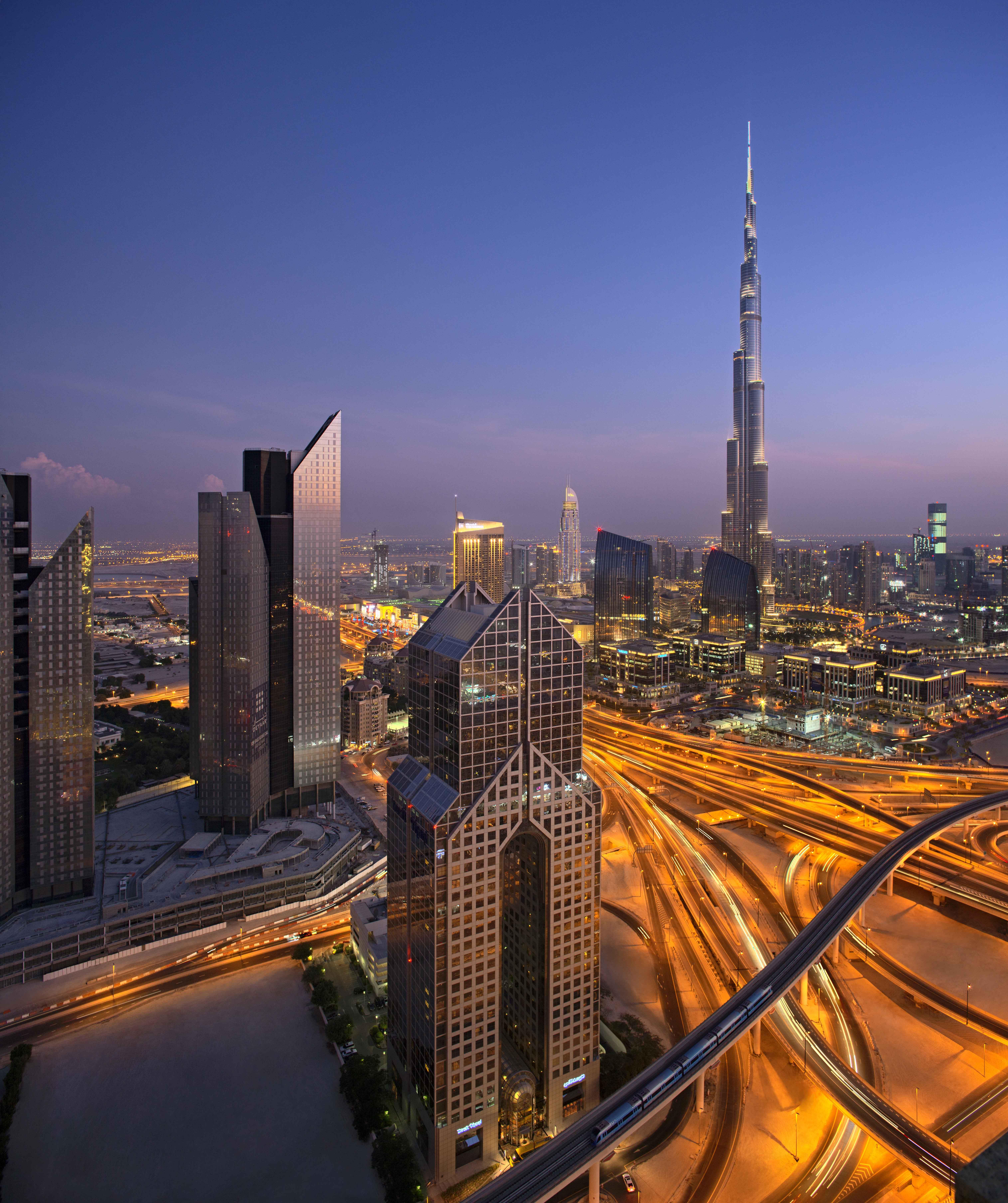 Dusit Thani Dubai Exterior photo The photo depicts a vibrant cityscape at dusk, featuring a mix of modern skyscrapers and distinctive architecture. Prominently, the Burj Khalifa, the tallest building in the world, rises majestically into the sky. The scene showcases intricate roadwa