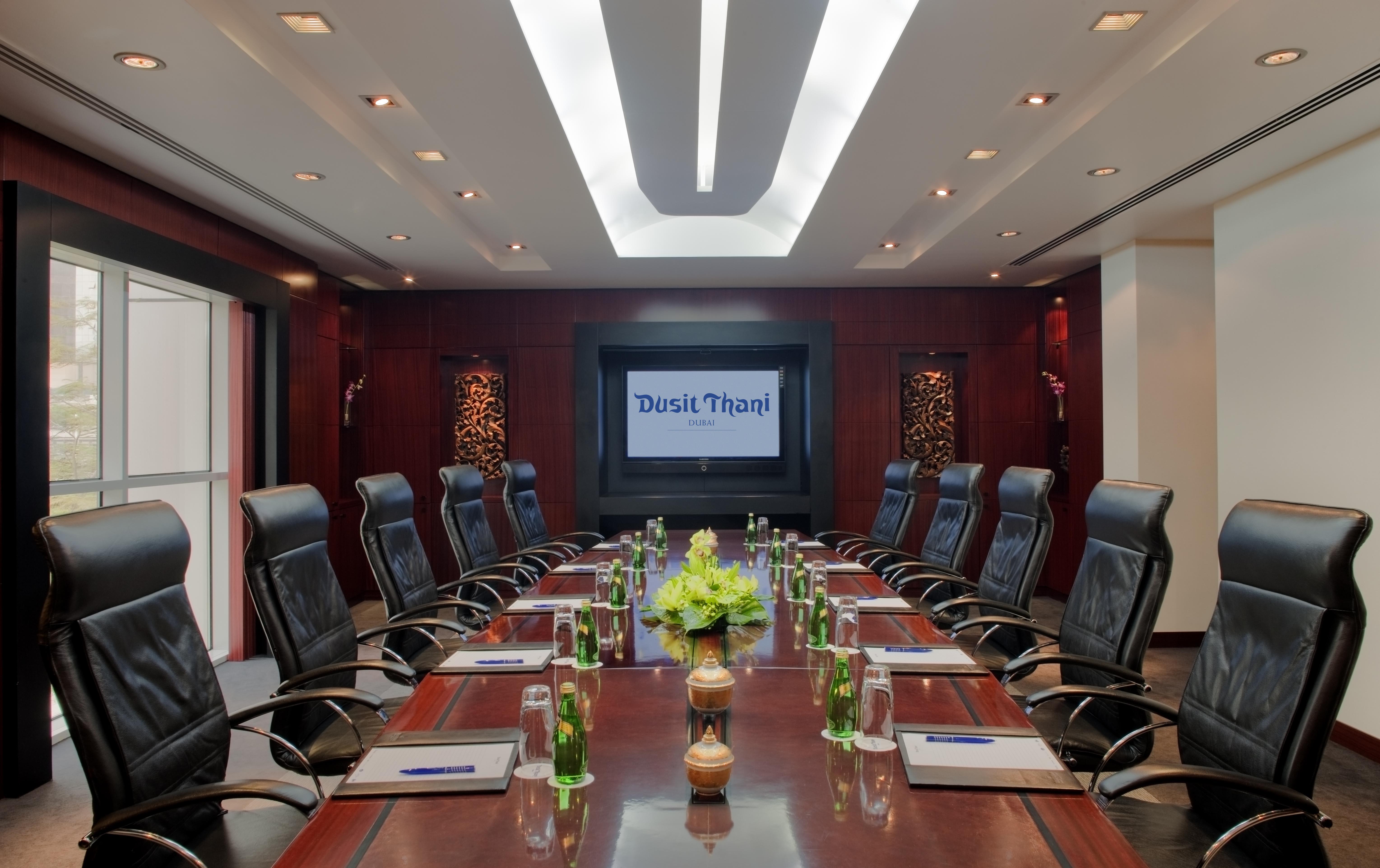 Dusit Thani Dubai Exterior photo The photo shows a sleek and modern conference room. It features a long wooden table surrounded by black leather executive chairs. At the center of the table, there is a decorative arrangement of green plants. On the table, there appear to be glass bo