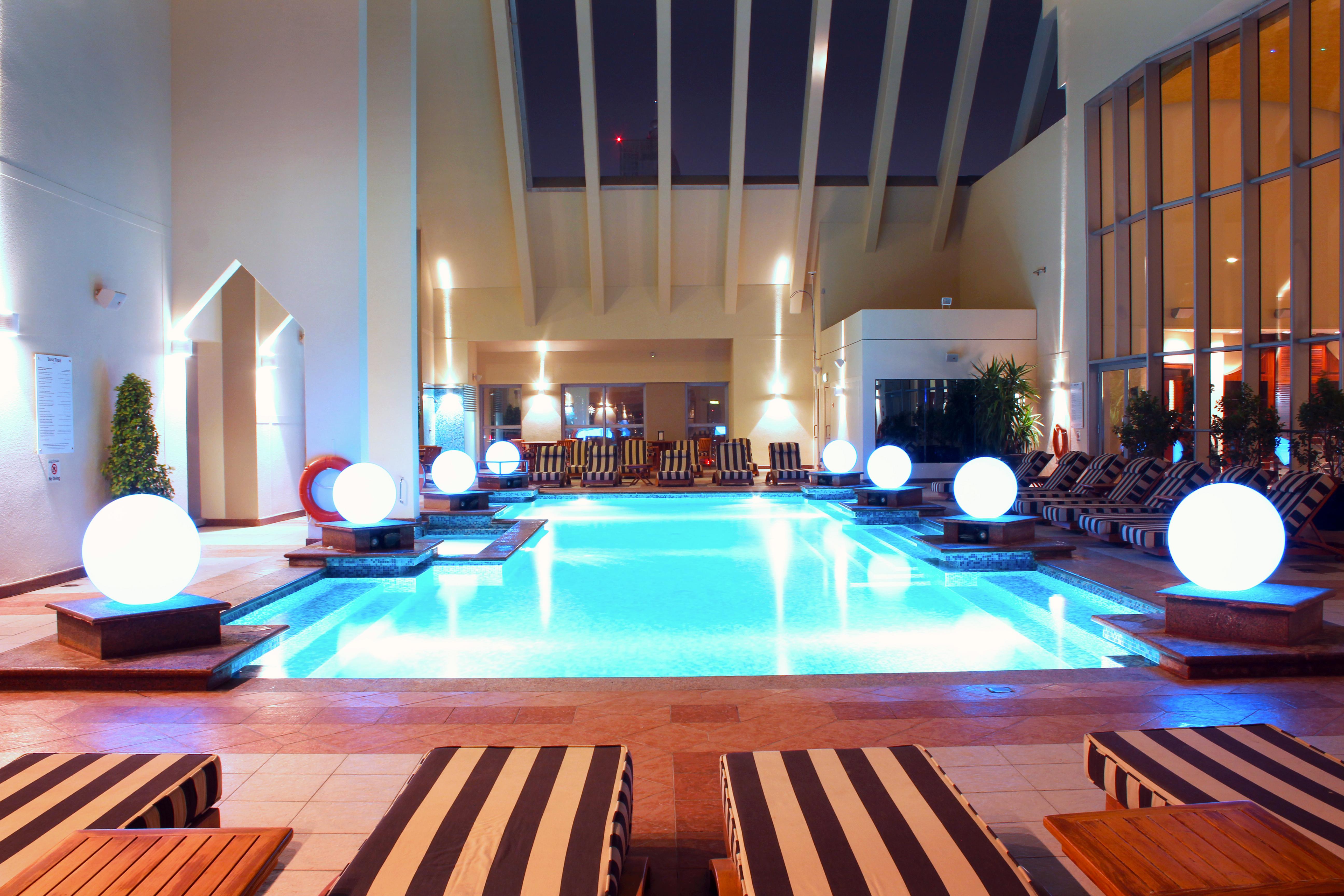 Dusit Thani Dubai Exterior photo The image shows an indoor swimming pool area. The pool is illuminated with soft blue lights, giving it a calm and inviting atmosphere. Surrounding the pool are several lounge chairs with striped cushions, positioned for relaxation. Large, glowing sph
