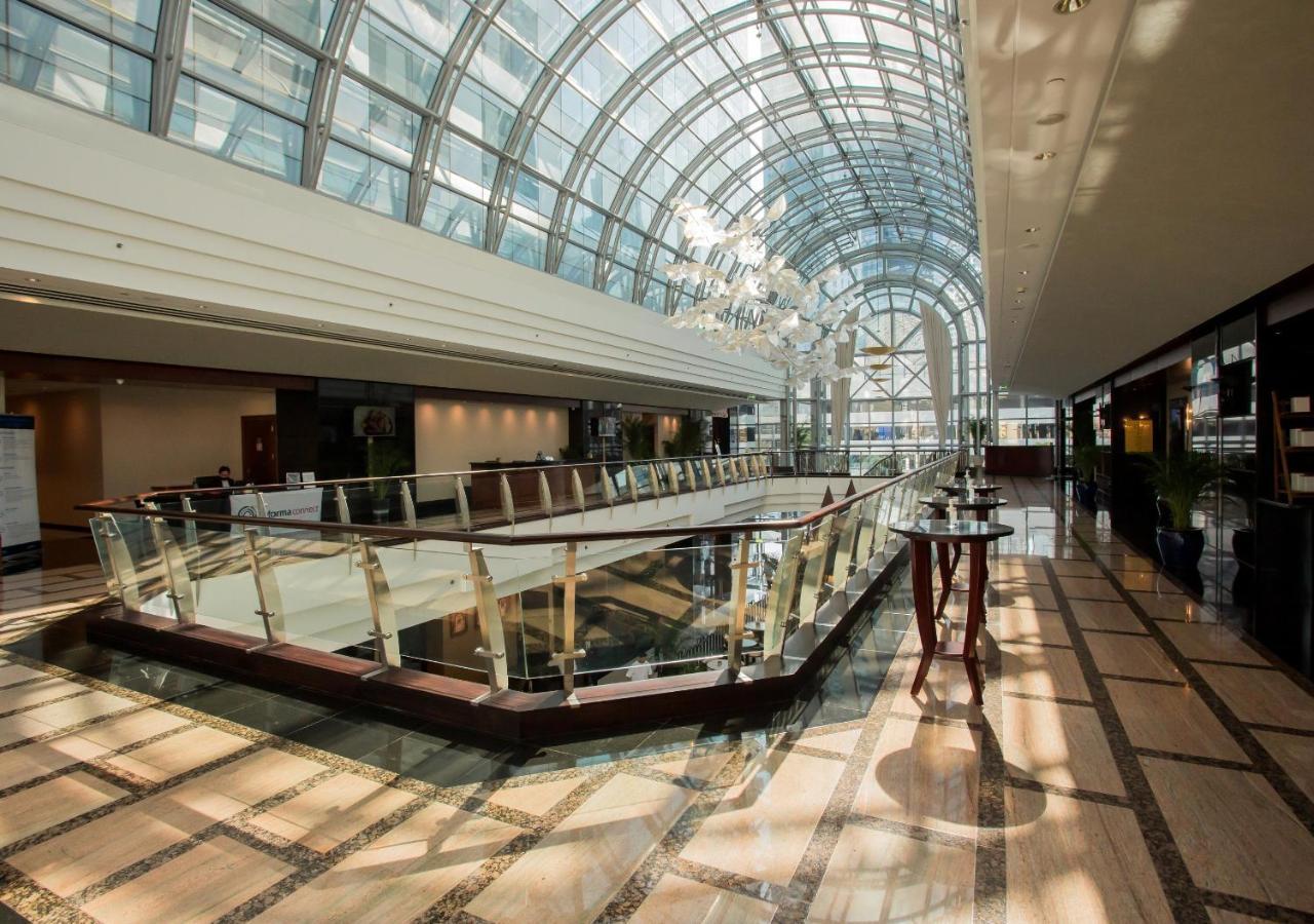 Dusit Thani Dubai Exterior photo The photo shows a spacious and modern interior, likely of a hotel or a shopping center. There is a large, glass dome overhead that allows natural light to flood the area, enhancing the open feel. The flooring is made of polished marble tiles, featuri