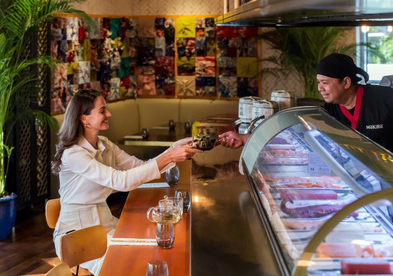 Dusit Thani Dubai Exterior photo The photo depicts a scene inside a cafe or ice cream shop. A woman is seated at a counter, smiling and reaching out to receive a drink or dessert from a staff member, who is standing behind the counter. The staff member appears friendly and is handin