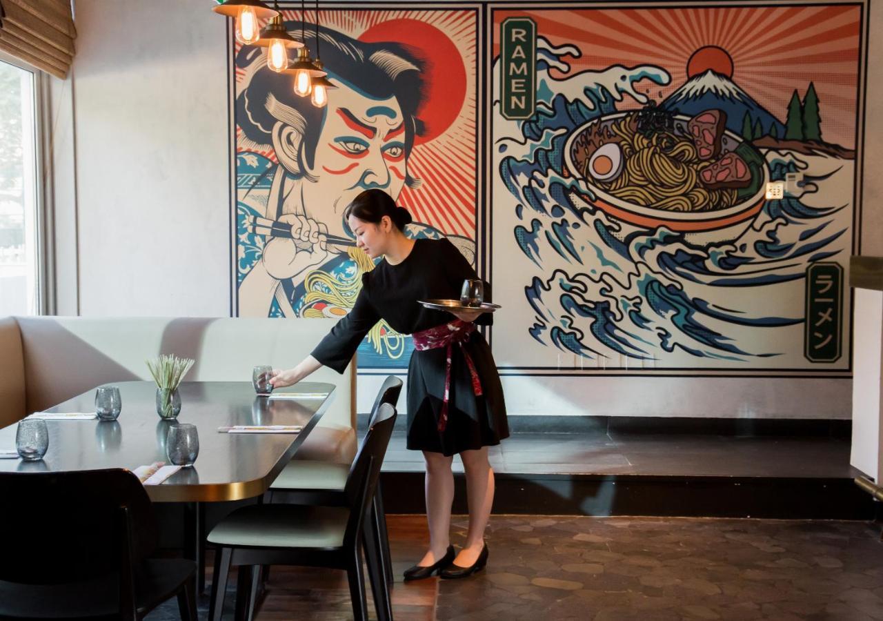 Dusit Thani Dubai Exterior photo The photo shows a woman in a black dress with a colorful sash, serving at a restaurant. She is placing a glass on the table while standing in front of a stylish interior. The walls are adorned with vibrant, graphic murals. One mural features a tradit