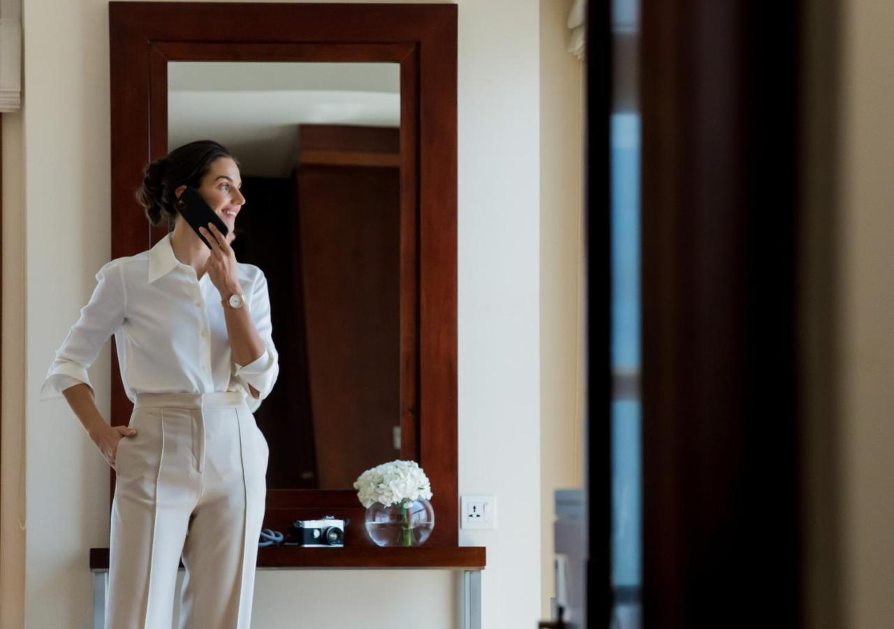 Dusit Thani Dubai Exterior photo The photo shows a woman dressed in a stylish, light-colored outfit. She is wearing a fitted white blouse and high-waisted trousers, and she appears to be smiling while holding her phone to her ear. The background features a large mirror and a small t