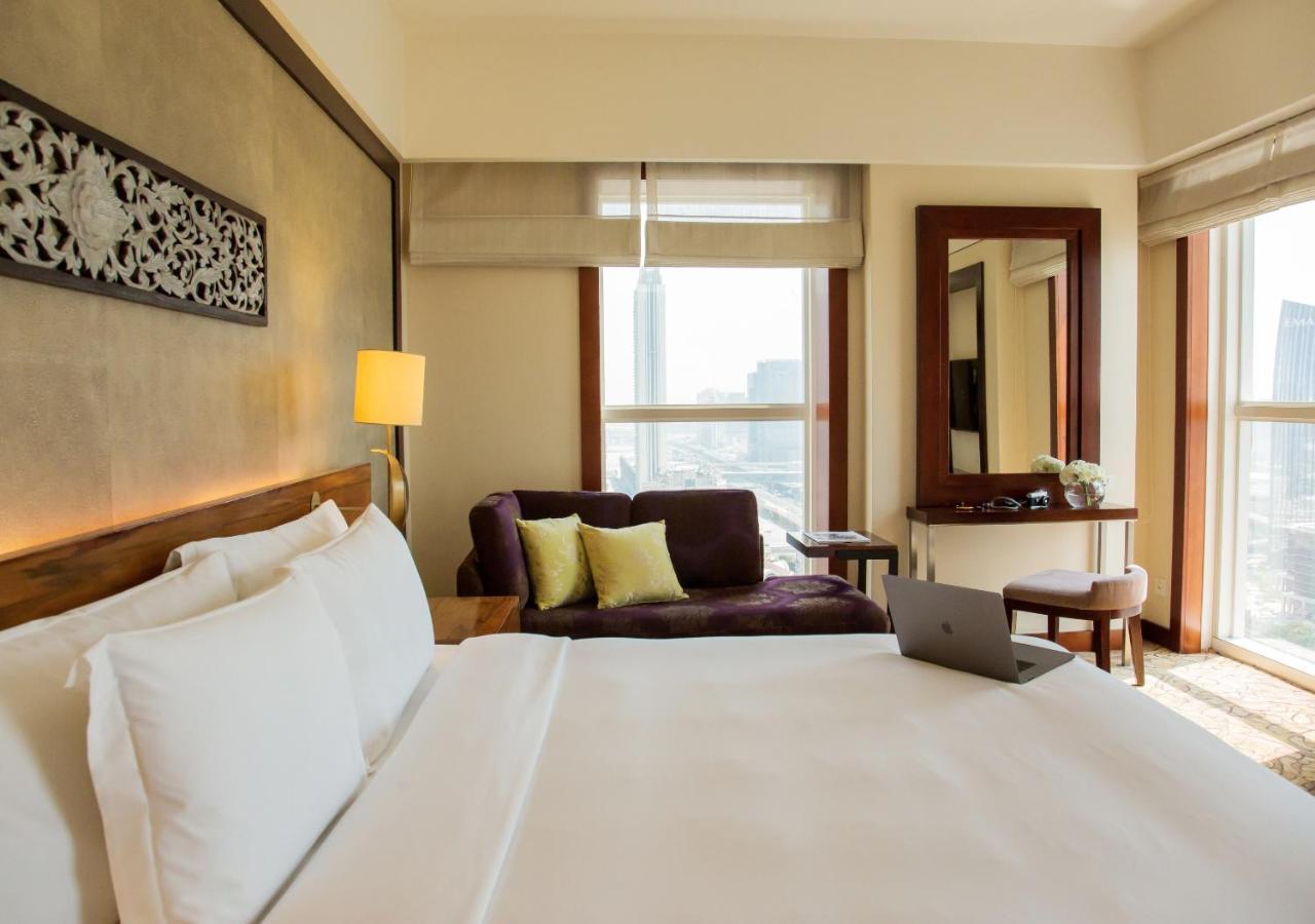 Dusit Thani Dubai Exterior photo The photo shows a well-furnished hotel room. In the foreground, there is a large bed with white linens. On the left side, a bedside lamp provides soft lighting. To the right, there is a cozy seating area with a dark purple couch and two green cushion