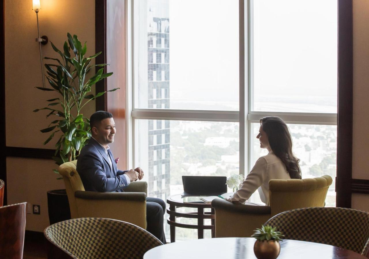 Dusit Thani Dubai Exterior photo The photo depicts two individuals seated in a modern, upscale setting, likely a coffee shop or lounge area. They are engaged in conversation, facing each other, with a small table between them. Natural light is coming through large windows, showcasin
