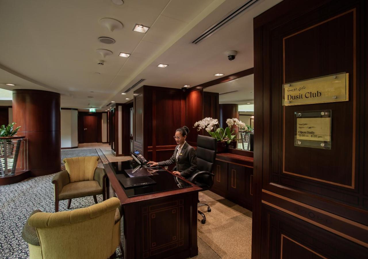 Dusit Thani Dubai Exterior photo The photo shows an elegant reception area within a hotel or a similar venue. A receptionist is seated at a wooden front desk, working on a computer. The desk is adorned with a floral arrangement, likely orchids, which adds a touch of sophistication t