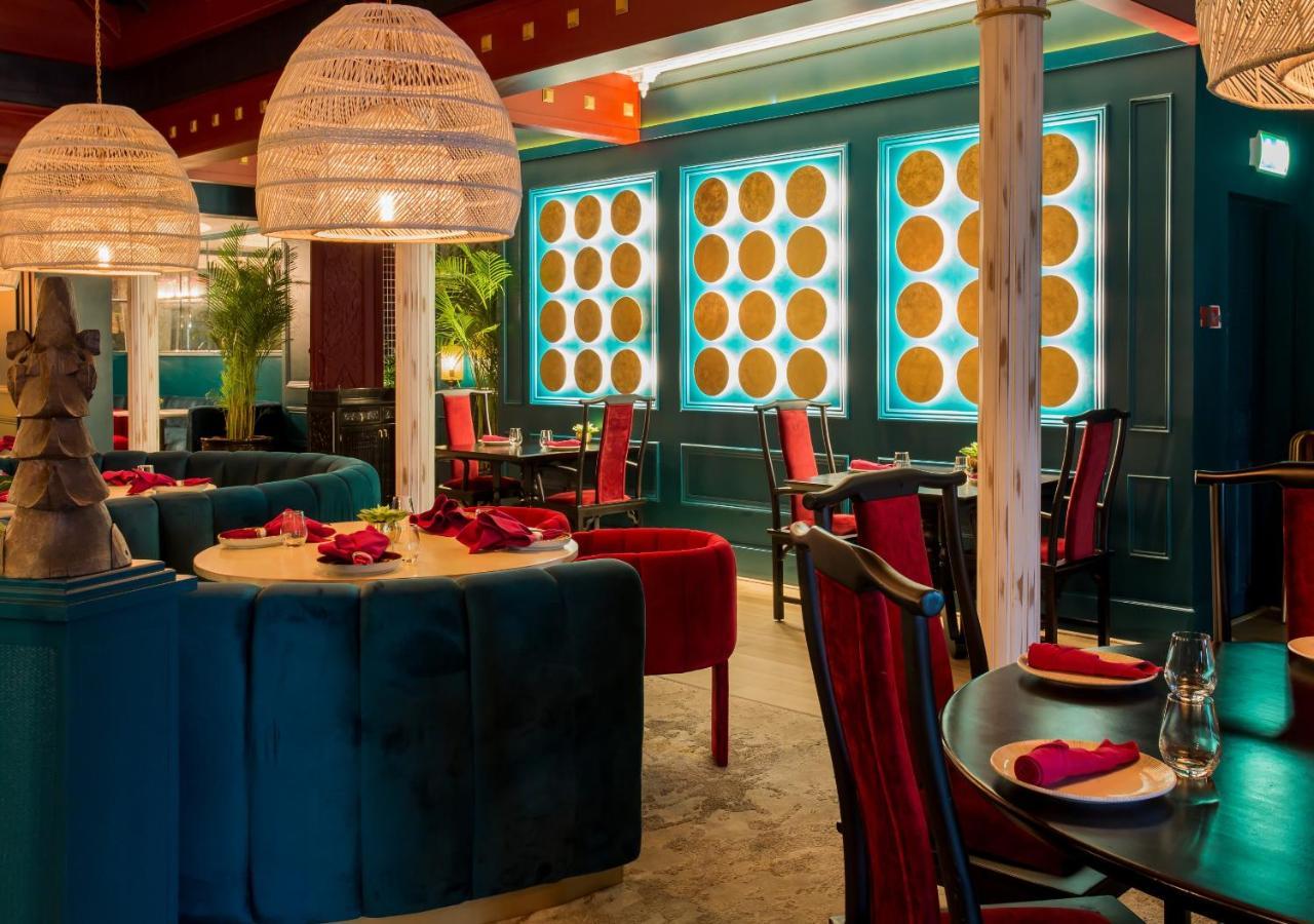 Dusit Thani Dubai Exterior photo The photo shows the interior of a stylish restaurant. The decor features a mix of vibrant colors, including rich blues and reds. There are plush seating areas, with some booths upholstered in dark blue fabric and chairs in a bold red. The lighting is