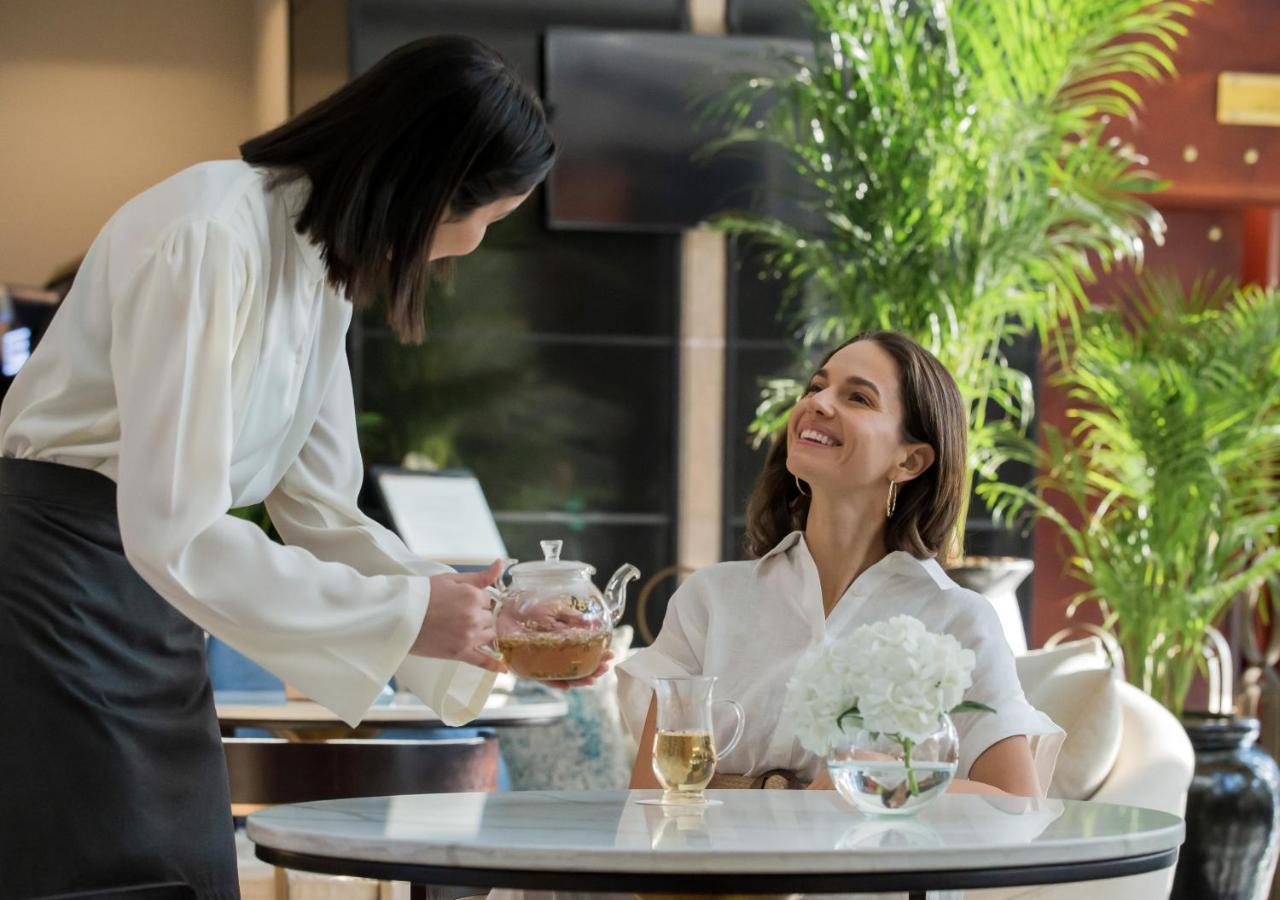 Dusit Thani Dubai Exterior photo The image depicts a scene in a sophisticated setting, likely a hotel lobby or a cafe. A smiling woman with long hair is seated at a table, dressed in a white outfit. She appears to be enjoying a pleasant conversation. A server, also dressed in formal