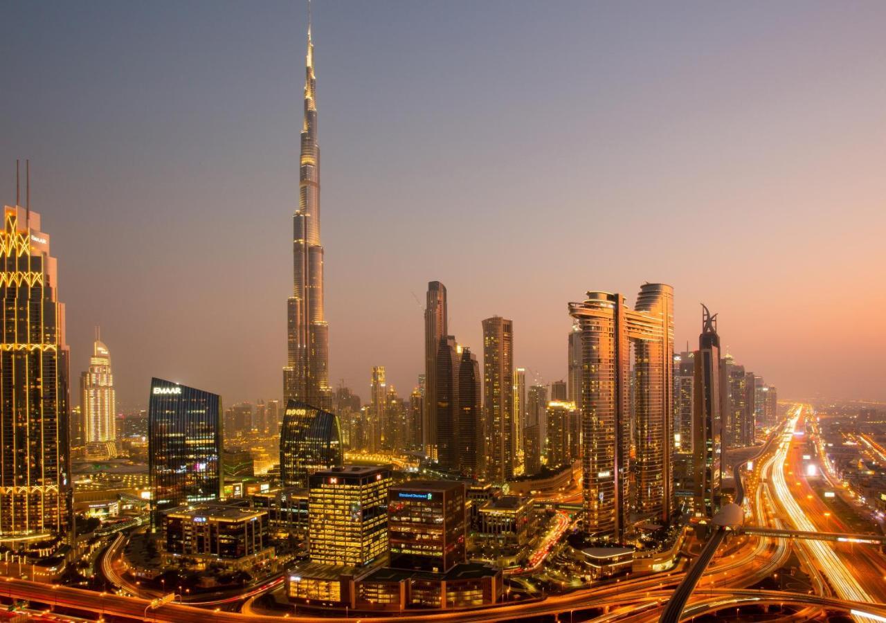 Dusit Thani Dubai Exterior photo The photo shows a vibrant skyline of a modern city at dusk, featuring tall skyscrapers bathed in the warm glow of the setting sun. Prominently in the background is a distinctive tall building, which is likely the Burj Khalifa, one of the tallest stru