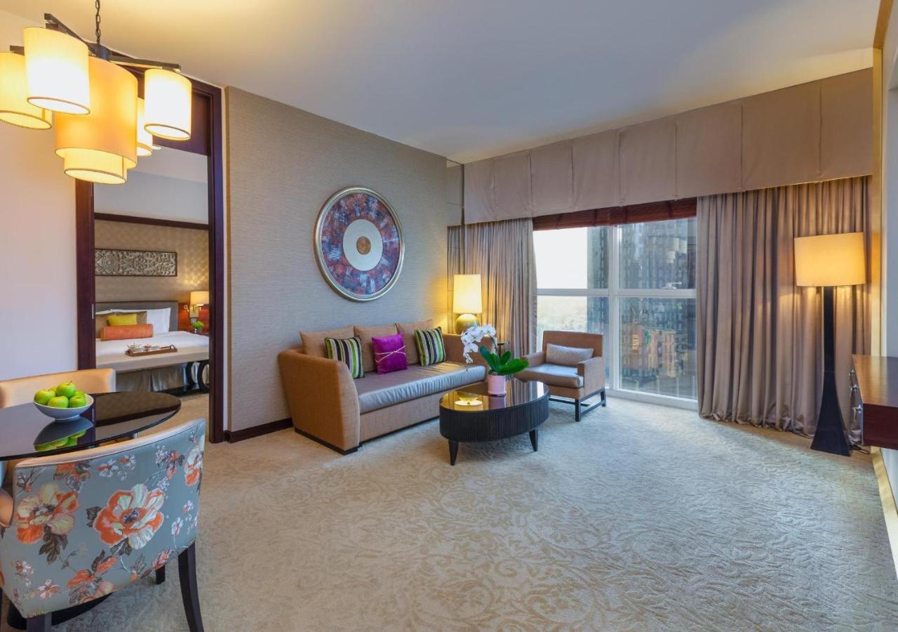 Dusit Thani Dubai Exterior photo The photo displays a modern hotel room or suite. It features a cozy seating area with a sofa adorned with colorful throw pillows. There’s a round coffee table in front of the sofa and a couple of chairs nearby. A large window lets in natural light, a