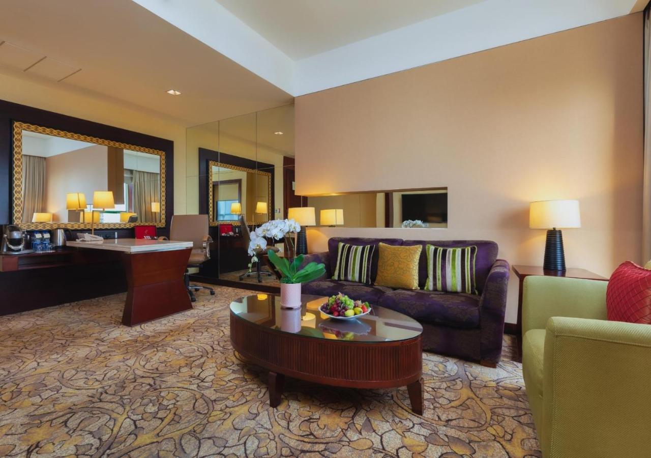Dusit Thani Dubai Exterior photo The photo depicts a stylish and well-furnished hotel suite. It features a cozy seating area with a dark purple sofa adorned with colorful cushions, accompanied by a light green armchair. There is a round coffee table in front of the sofa, topped with
