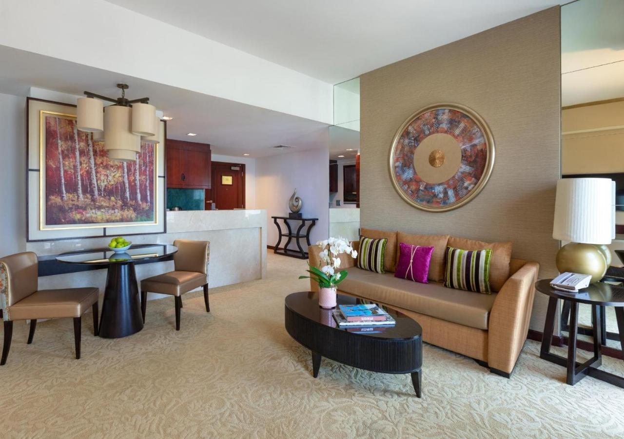 Dusit Thani Dubai Exterior photo The photo shows a modern and stylish living area. It features a comfortable brown sofa with decorative pillows in various colors. In front of the sofa is a round coffee table with some books and a vase with flowers on it. To the left, there is a dini