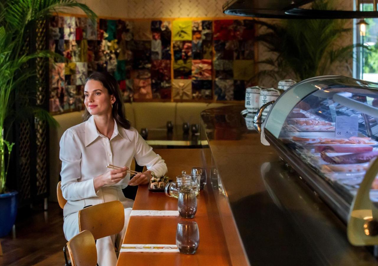 Dusit Thani Dubai Exterior photo The photo shows a woman sitting at a counter in a stylish café or restaurant. She appears to be enjoying a moment of relaxation, wearing a light-colored outfit. The setting features vibrant and artistic decor in the background, with a variety of colo
