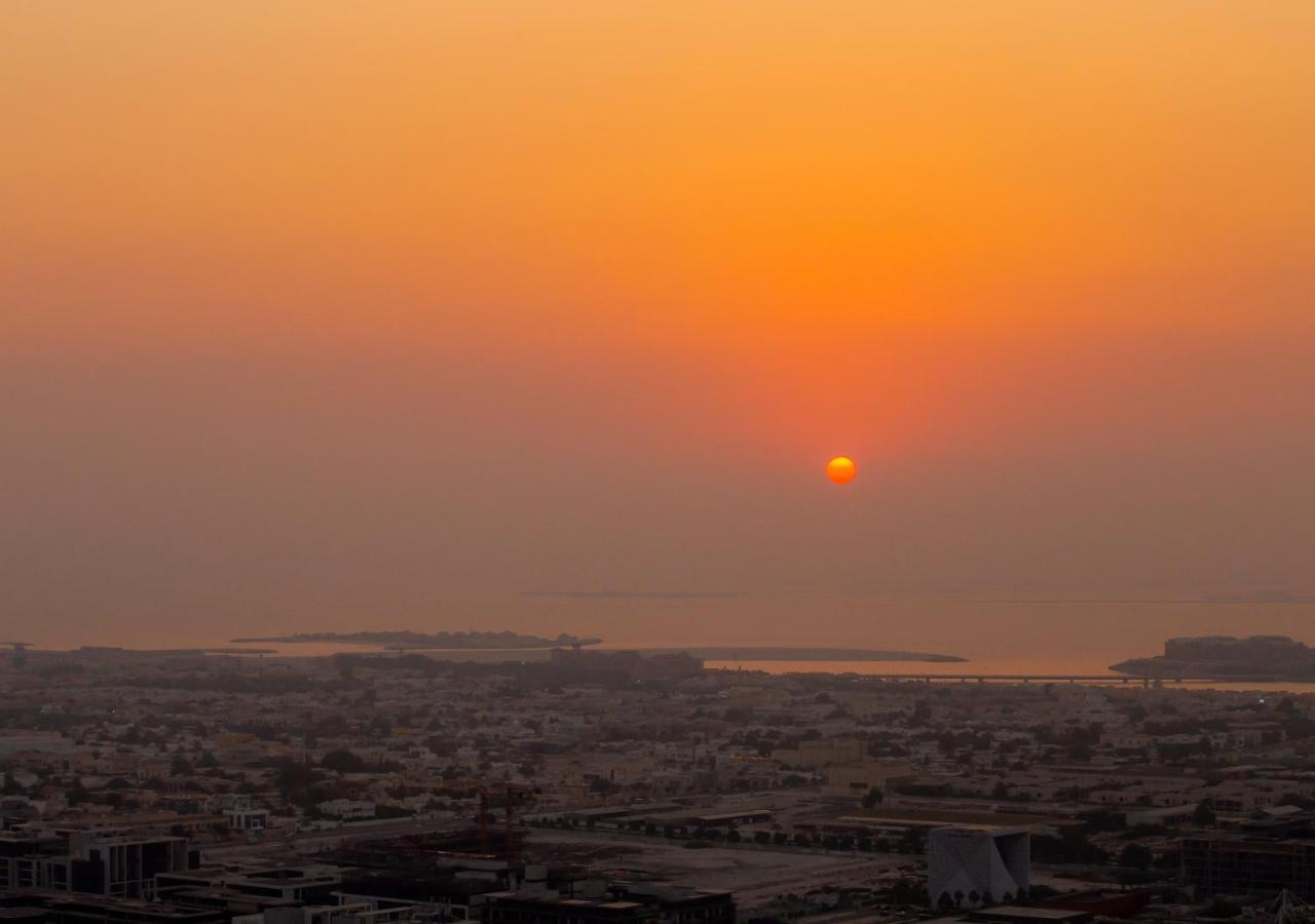Dusit Thani Dubai Exterior photo The photo shows a beautiful sunset over a cityscape. The sun is depicted as a small, glowing orb on the horizon, surrounded by a gradient of warm colors, ranging from deep orange to soft yellow. The foreground features a sprawling urban area with bui