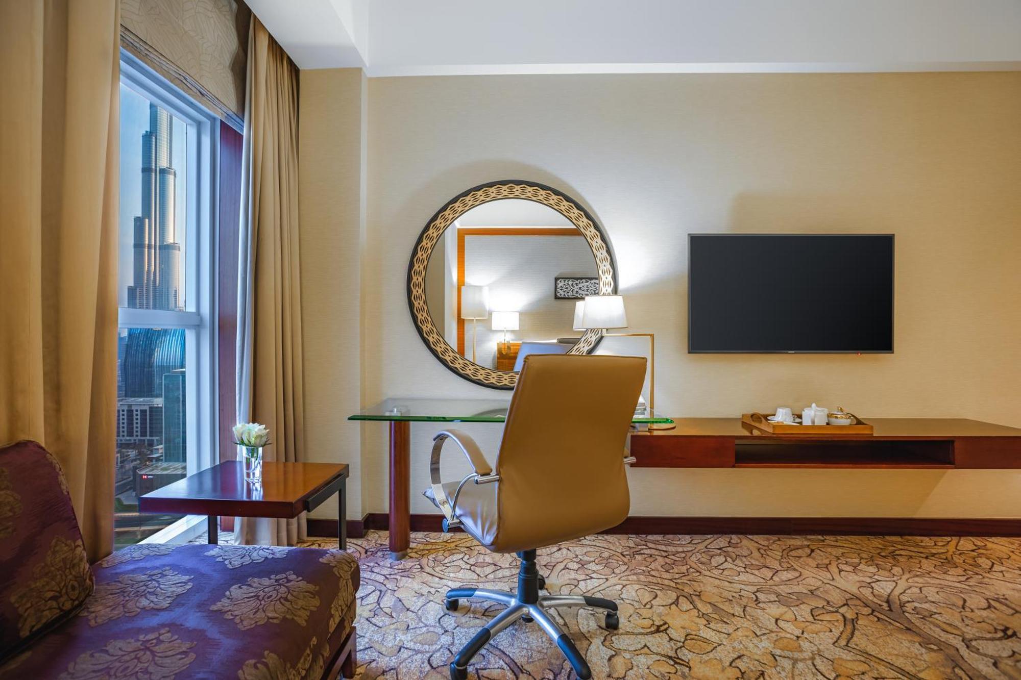 Dusit Thani Dubai Exterior photo The image shows a hotel room with a modern and elegant design. There is a comfortable chair and a small desk with a glass top, where a decorative item and a lamp are placed. A large round mirror is mounted on the wall above the desk, reflecting some 