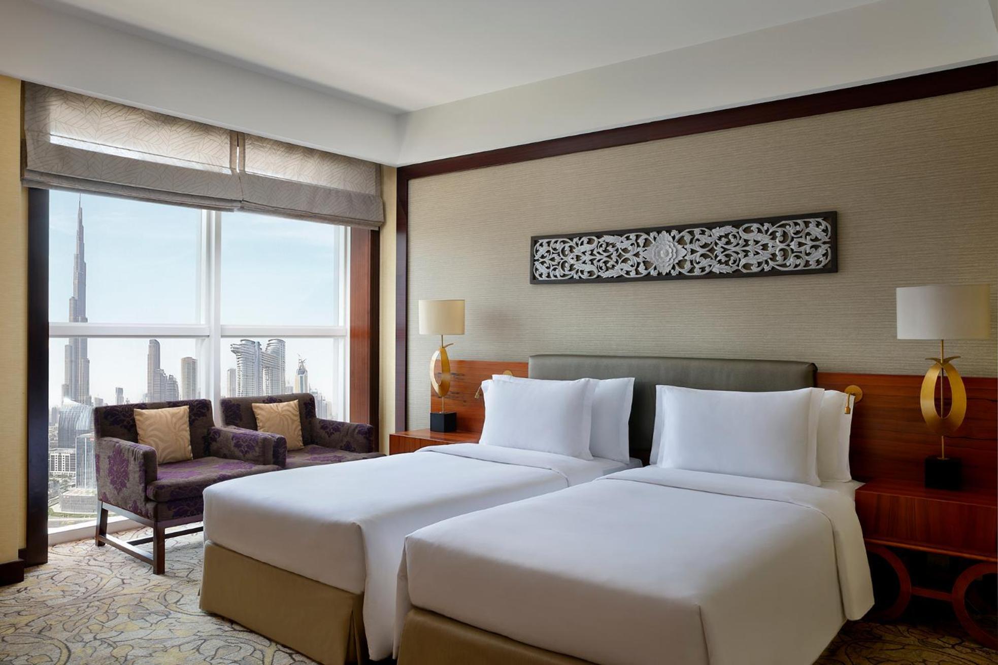 Dusit Thani Dubai Exterior photo The photo shows a well-decorated hotel room featuring two twin beds with white linens. There is a modern design aesthetic, with a light-colored wall and a decorative piece above the beds. The room has a large window that offers a view of the city, in