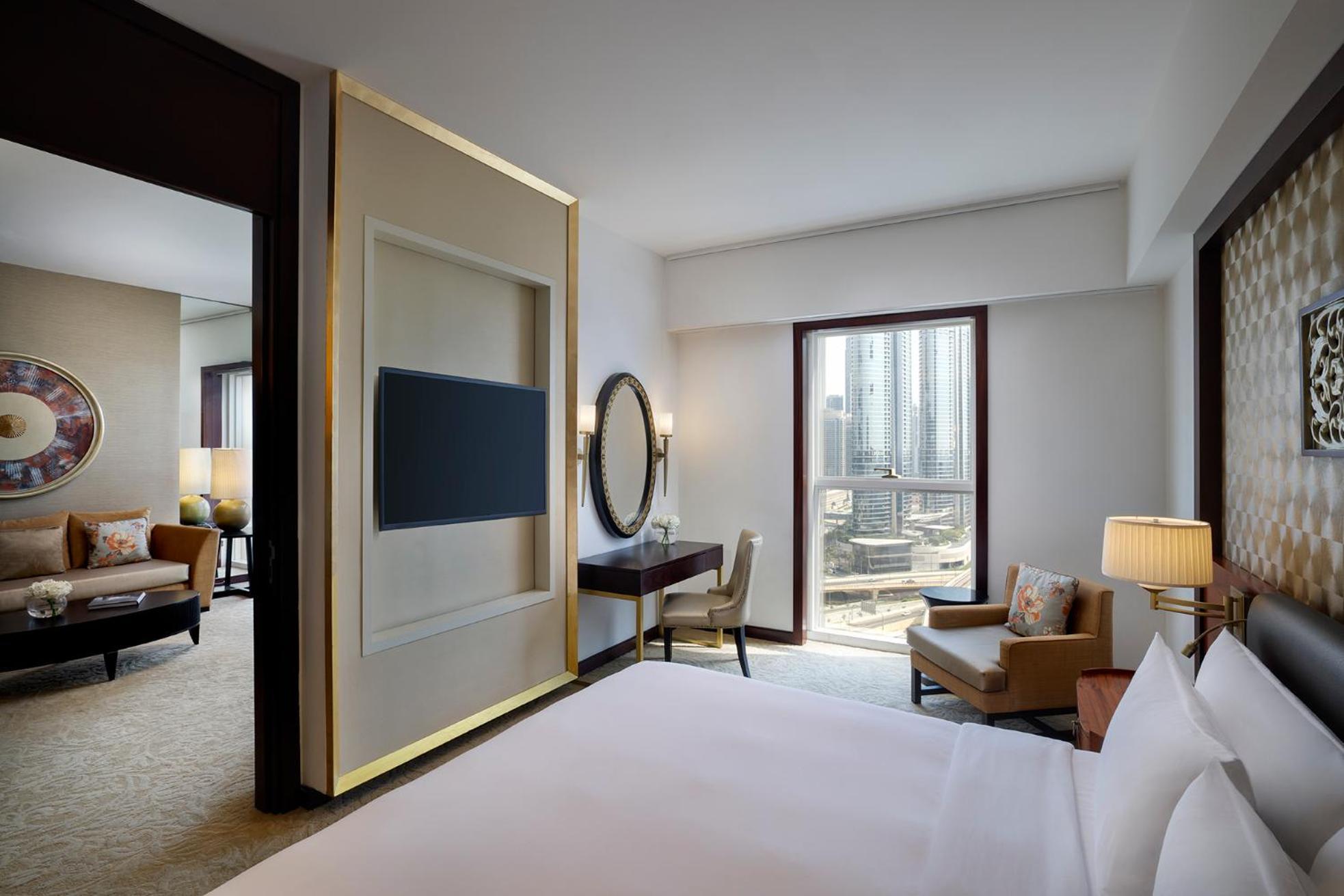 Dusit Thani Dubai Exterior photo The photo shows a modern hotel room featuring a comfortable bed with white linens in the foreground. Behind the bed, there is a stylish partition with a wall-mounted TV. To the side, a desk with a circular mirror is visible, accompanied by a chair. A