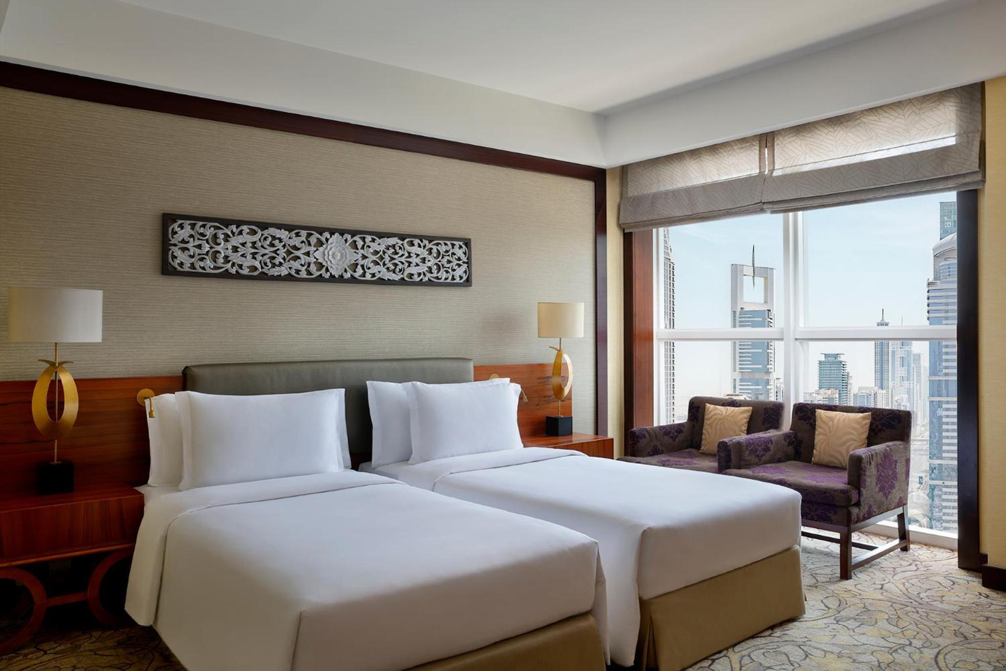Dusit Thani Dubai Exterior photo The photo shows a hotel room featuring two twin beds with white bedding. The room has a modern and elegant design, with a wooden headboard and decorative panel above the beds. There's a seating area with two purple chairs and a small table next to a 
