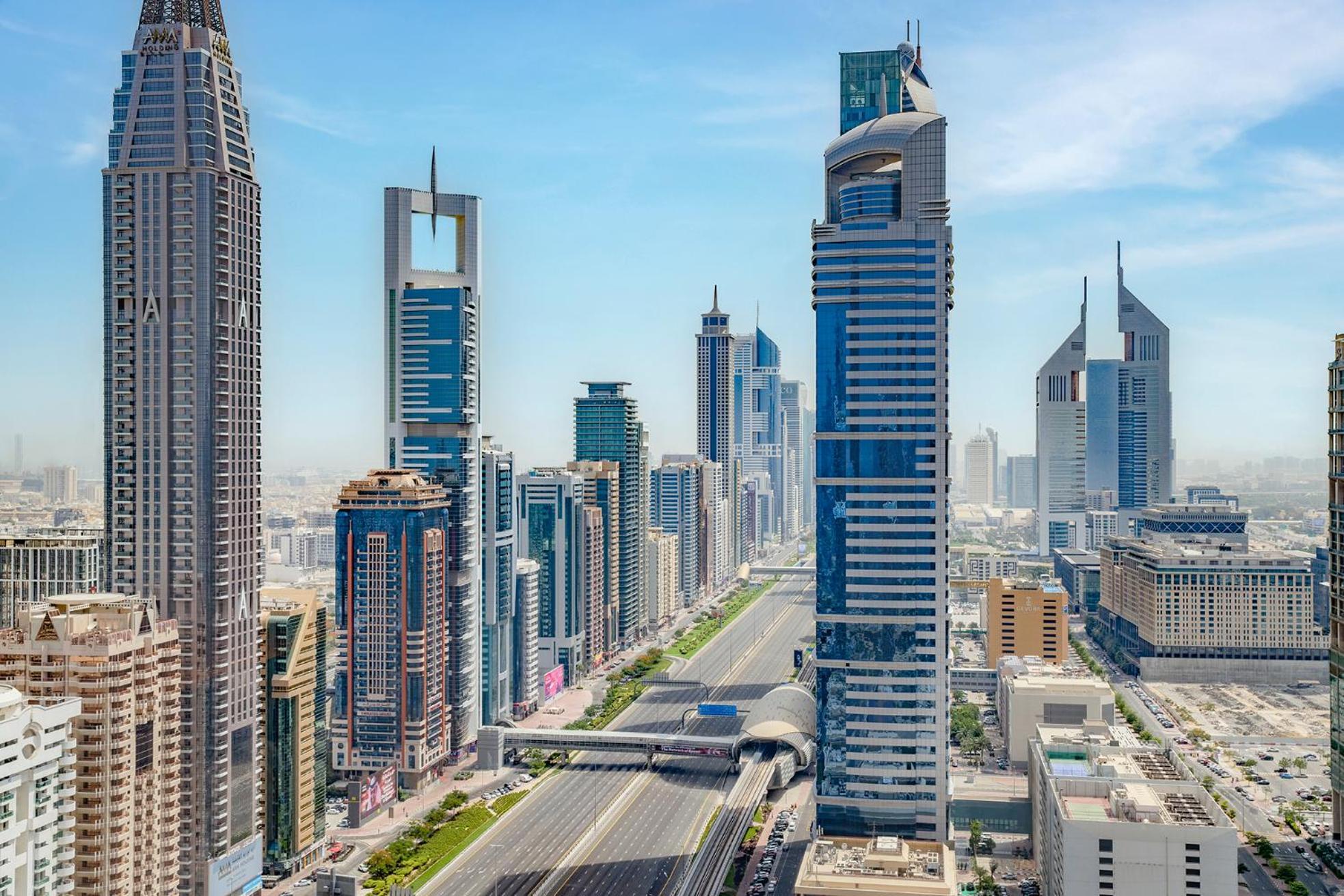 Dusit Thani Dubai Exterior photo The photo depicts a vibrant urban skyline featuring a variety of modern skyscrapers. The buildings showcase diverse architectural styles, some with sleek glass facades and unique shapes. A wide road stretches through the foreground, indicative of a b