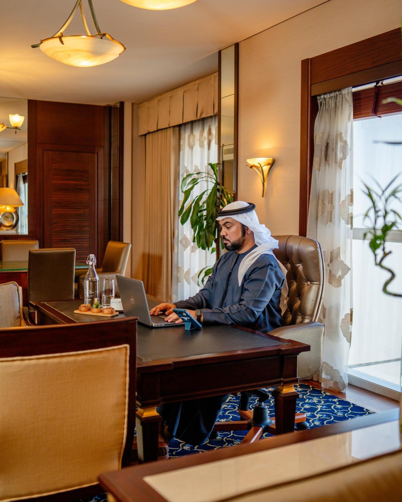 Dusit Thani Dubai Exterior photo The photo depicts a man seated at a desk in a well-furnished office environment. He is wearing traditional attire, including a kandura and a headscarf. The workspace features a modern laptop, and there are snacks or pastries on a small plate beside h