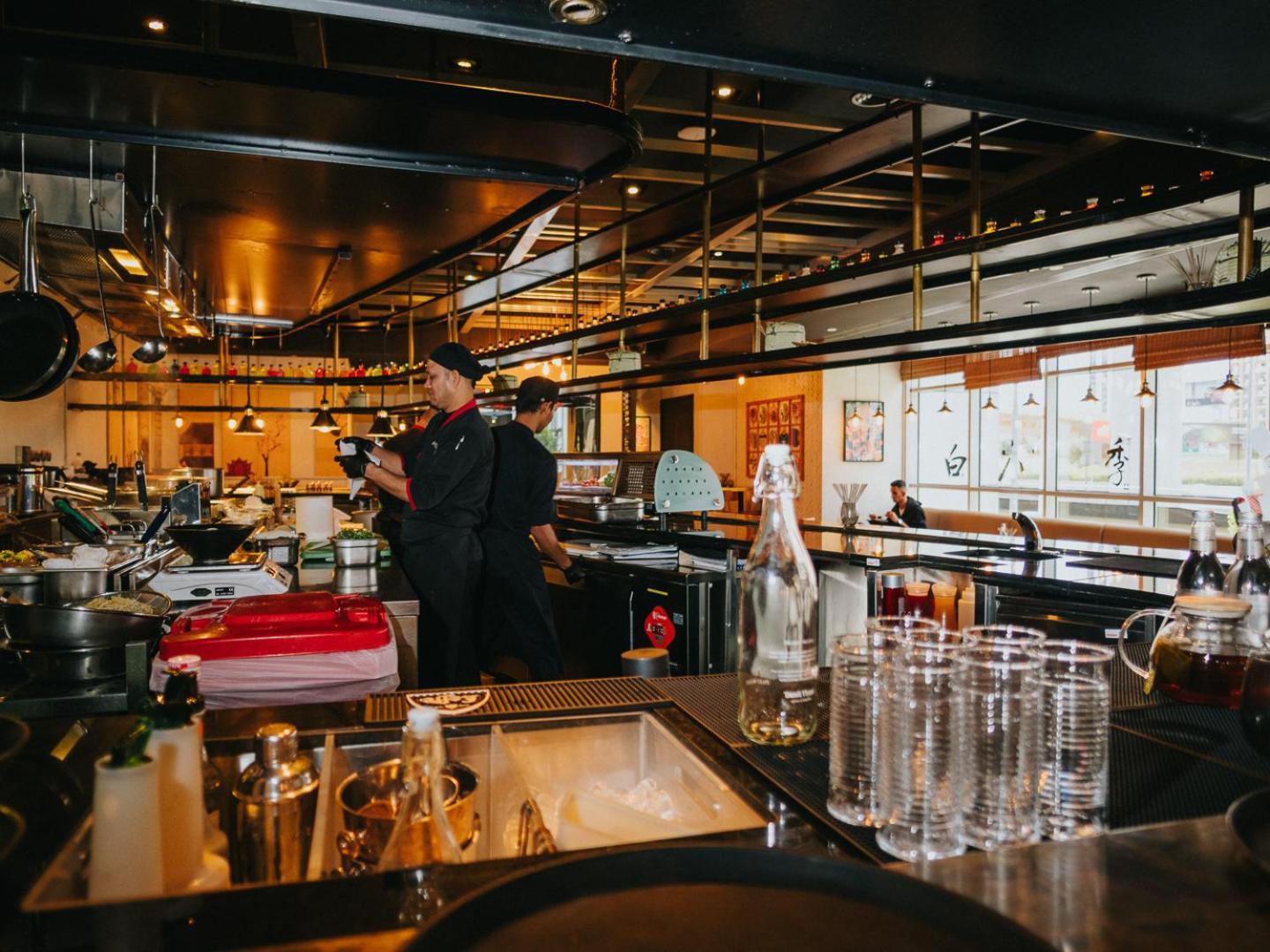 Dusit Thani Dubai Exterior photo The photo shows an interior view of a restaurant or café. In the foreground, there is a bar area with glasses and various utensils visible. Two staff members, dressed in black uniforms, are engaged in conversation or work. The background features an 
