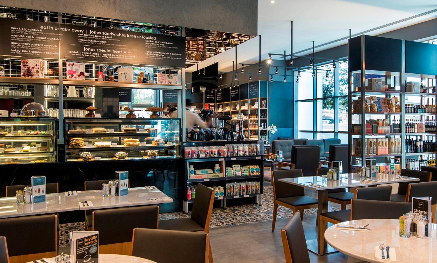 Dusit Thani Dubai Exterior photo The photo shows a modern café or restaurant interior. There are tables set for dining with simple yet stylish chairs. A glass display case filled with various food and beverage items is prominently featured, showcasing pastries and other offerings. I