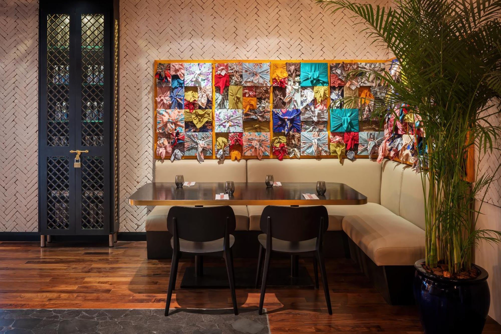 Dusit Thani Dubai Exterior photo The photo shows a cozy restaurant interior featuring a seating area with a large beige bench and a wooden table. Behind the seating area is a colorful mural made up of various textured and patterned fabrics or materials, creating a vibrant backdrop. 