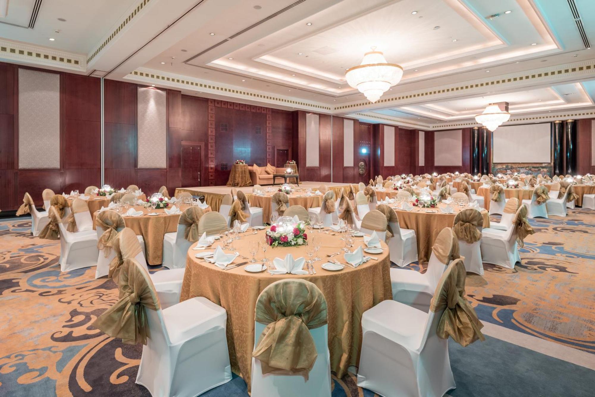 Dusit Thani Dubai Exterior photo The photo depicts an elegant banquet or event hall set up for a formal gathering. The room features multiple round tables, each adorned with gold tablecloths and surrounded by white chairs. The chairs are decorated with golden sashes. Centerpieces on
