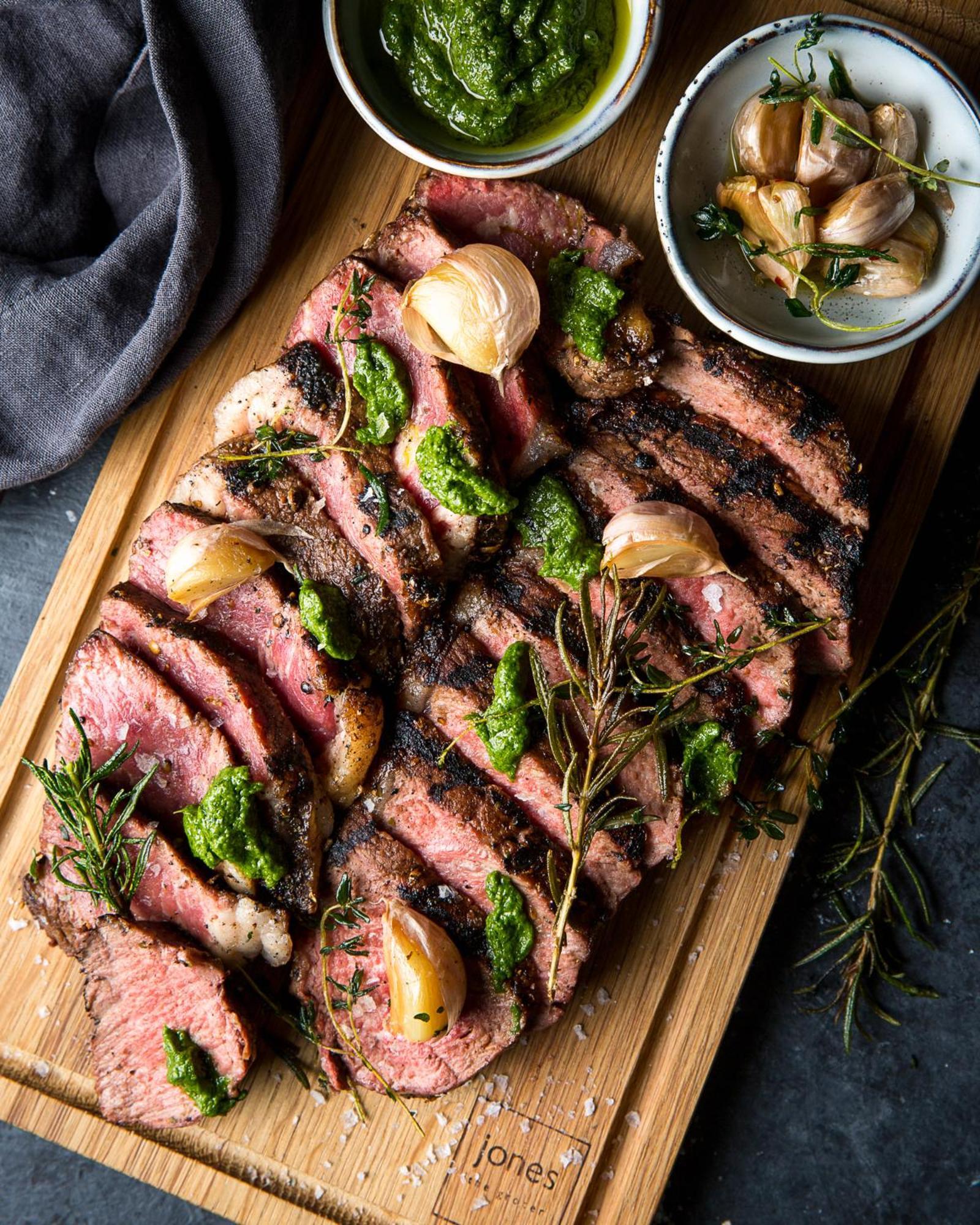 Dusit Thani Dubai Exterior photo The photo shows a beautifully arranged platter of grilled steak, sliced into portions. The steak has a rich, charred exterior and is garnished with green herb sauce, possibly pesto. There are roasted garlic cloves and sprigs of fresh herbs, like rose