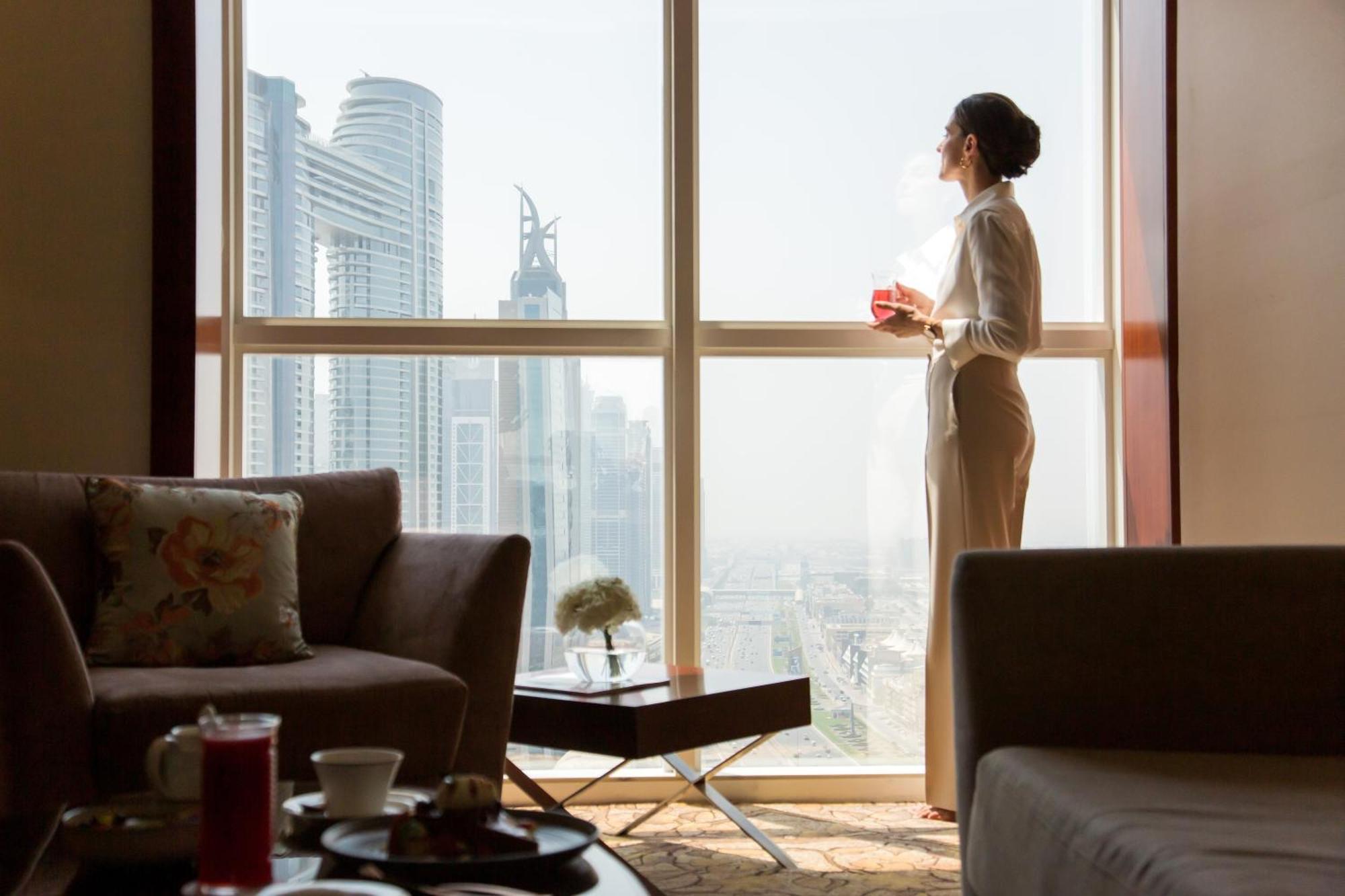 Dusit Thani Dubai Exterior photo The photo depicts a woman standing by a large window, looking out at a city skyline filled with modern skyscrapers. She appears to be in a well-furnished room, possibly a hotel or a luxurious apartment, with a stylish sofa and a coffee table in the f