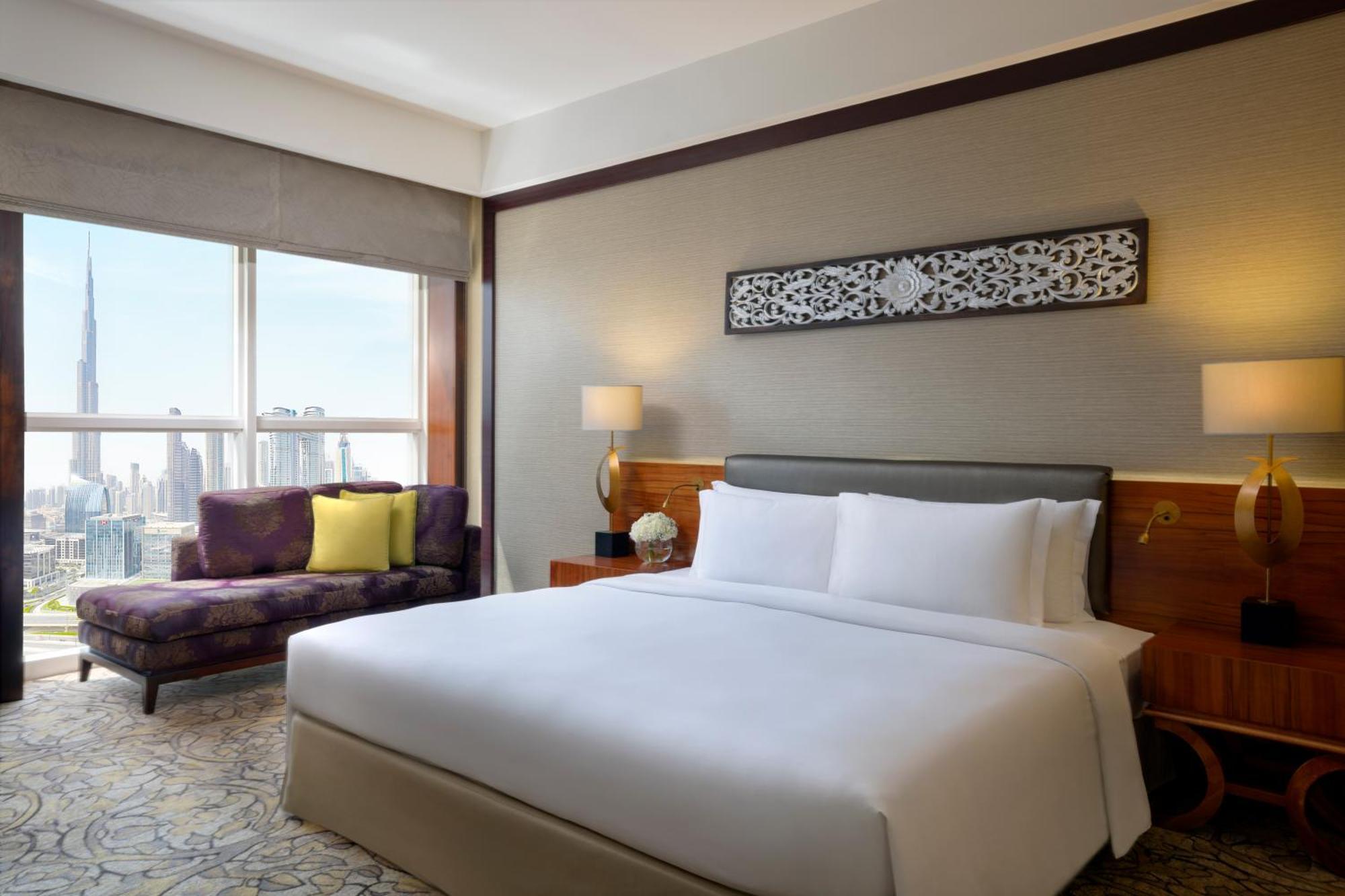 Dusit Thani Dubai Exterior photo The photo shows a modern hotel room featuring a large bed with white linens, a stylish headboard, and decorative pillows. To the left, there's a small sitting area with a purple couch and a decorative cushion, providing a contrast to the neutral tone