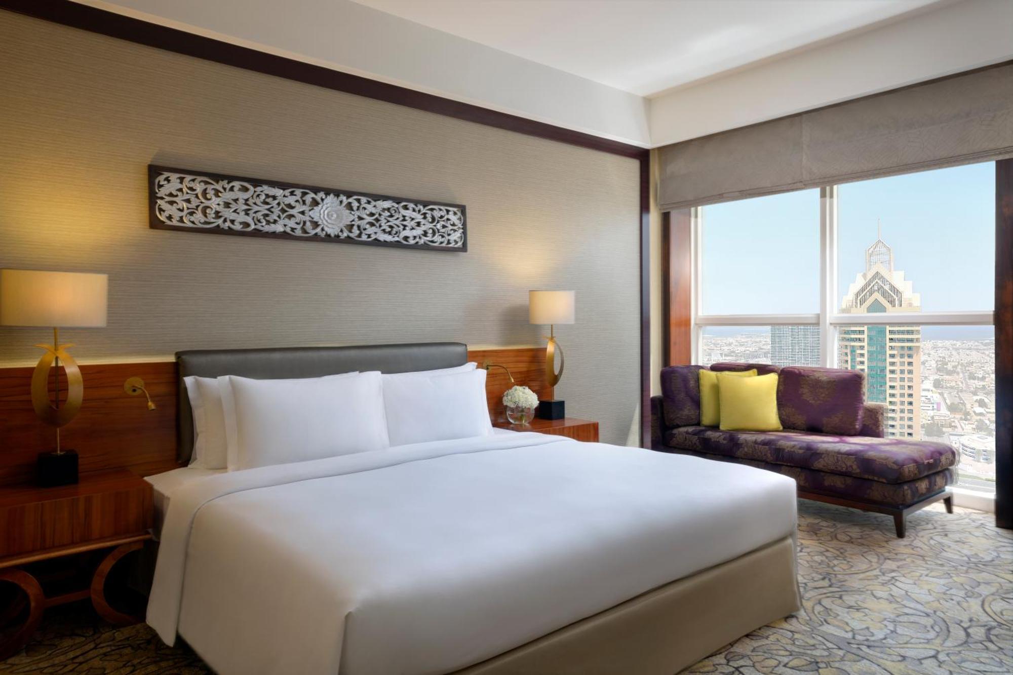Dusit Thani Dubai Exterior photo The photo shows a modern hotel room featuring a large bed with crisp white bedding. There are two bedside lamps on either side of the bed, and a decorative piece is hanging on the wall above it. In the corner, there is a comfortable chair next to a w